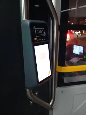 A contactless card validator at the door of a Craiova city bus, screen showing line E1R and the next stop