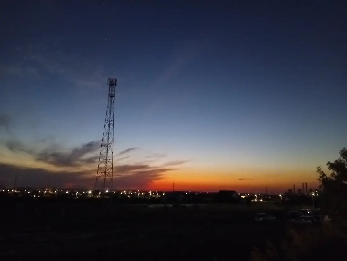 Sunset behind a telecoms mast, the same evening — chimney stacks on the skyline to the right, the city’s lights coming on below