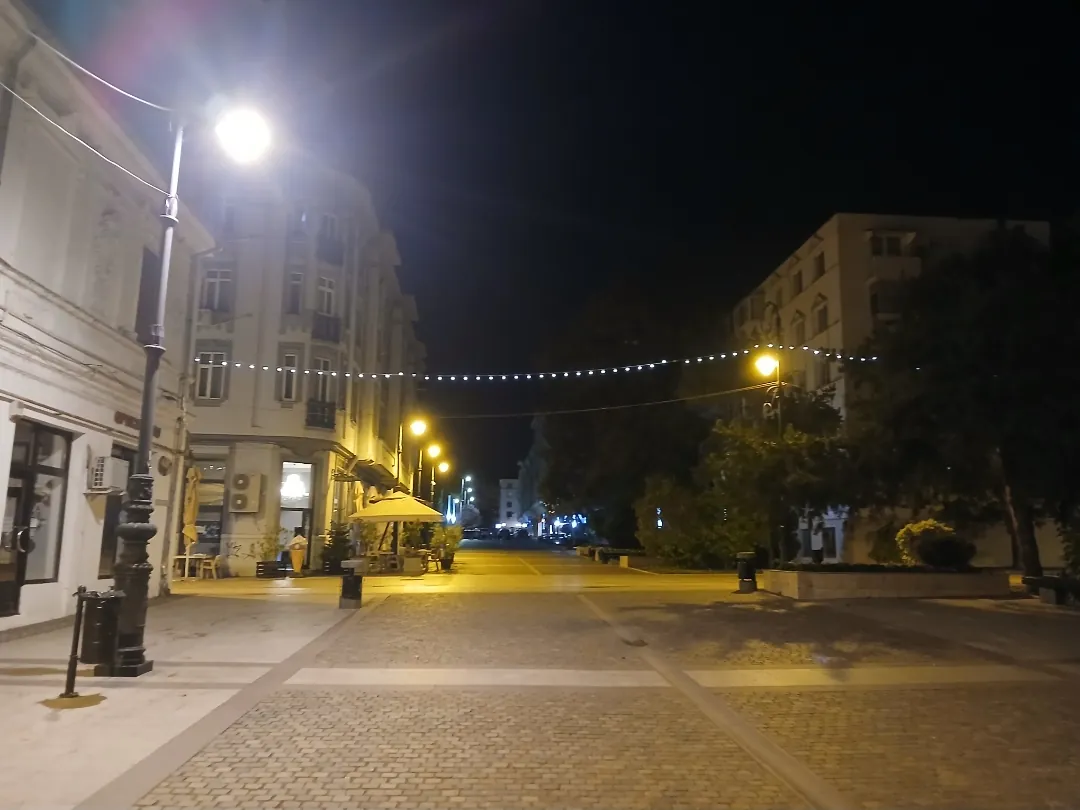 The old centre after dark — a pedestrian street under a string of lights, lamps on, one café umbrella still up. Street not yet named