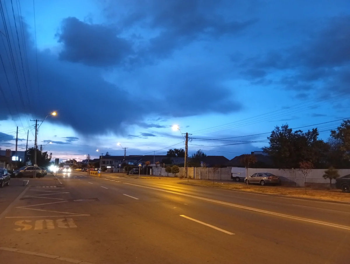 Dark storm clouds over a wide road on the edge of Craiova at dusk, streetlights just on and a bus stop in the foreground, 22 September 2026