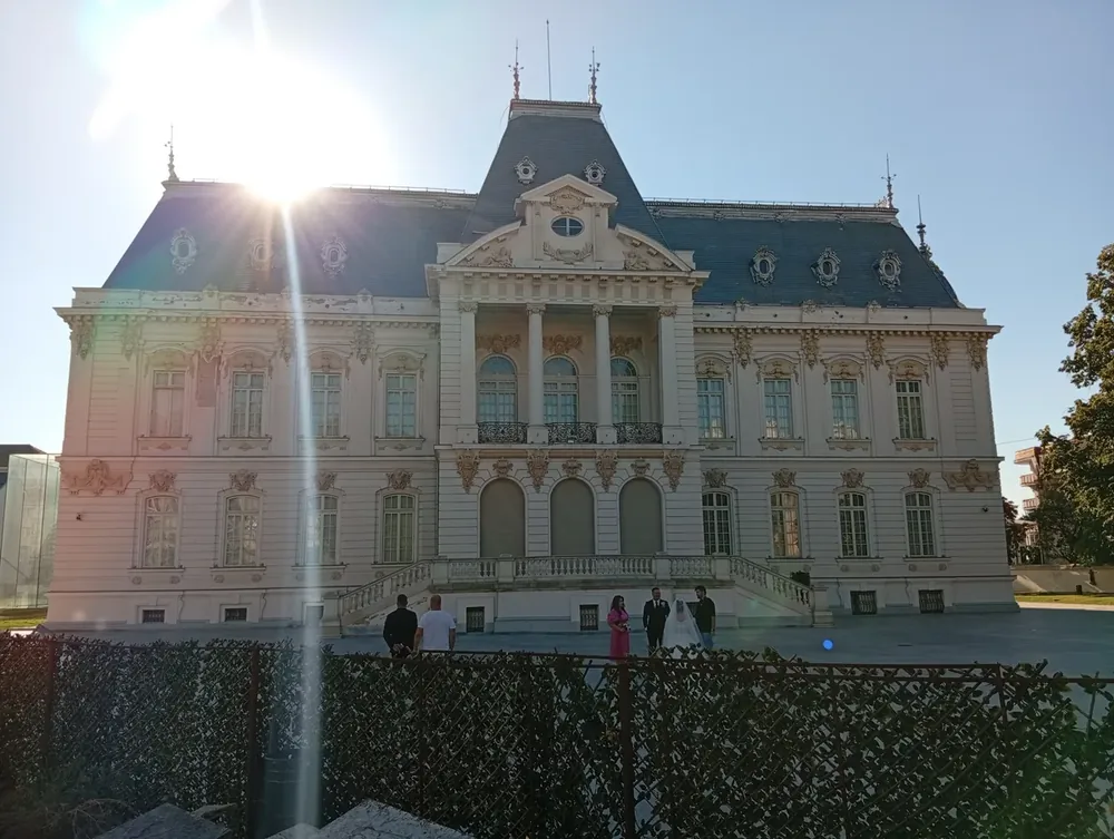 The Jean Mihail Palace in Craiova, home of the art museum — a French-baroque façade with a columned central bay and slate mansard roofs, a wedding party on the forecourt in low afternoon sun