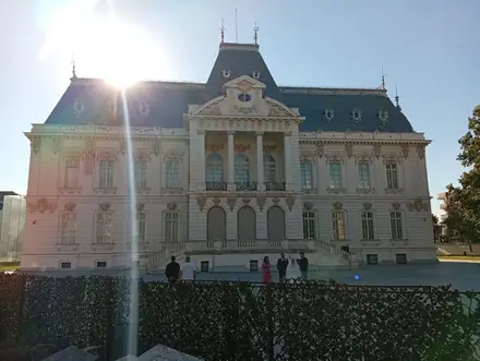 The Jean Mihail Palace in Craiova, home of the art museum — a French-baroque façade with a columned central bay and slate mansard roofs, a wedding party on the forecourt in low afternoon sun