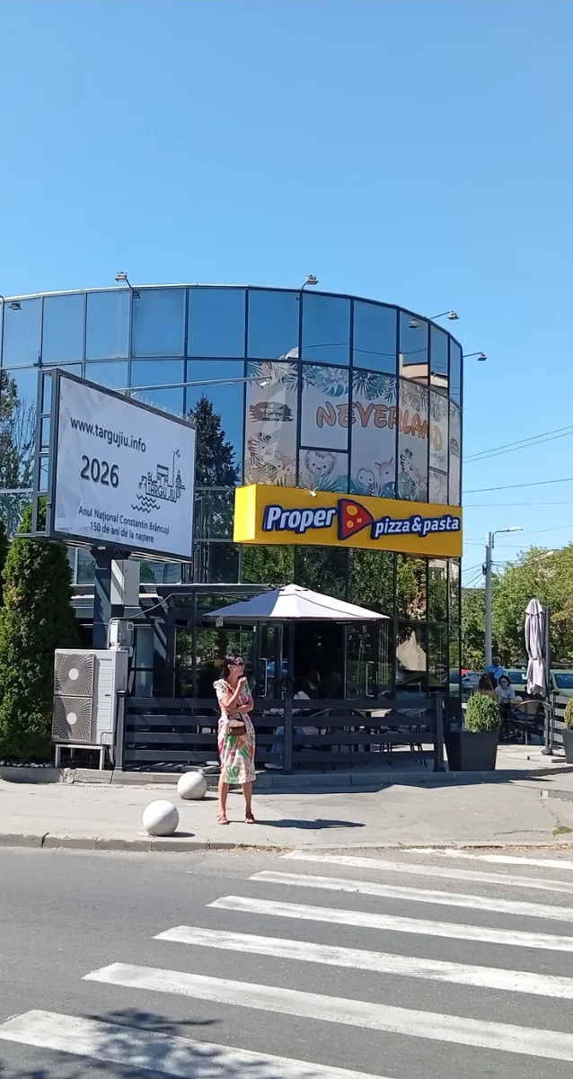 A curved glass drum on Strada Opanez, pizzeria and terrace on the ground floor and a children’s play centre painted across the glass above it. The billboard beside it is advertising Târgu Jiu — 2026 as the national Brâncuşi year, 150 years since his birth — and has nothing to do with the building