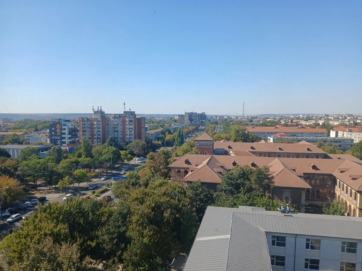 Craiova from a high-rise on a clear October afternoon: a tree-lined boulevard running straight to the horizon, blocks of flats on the left, a long red-tiled complex on the right, and the hills beyond the city on the skyline. The blue road sign below points to Timisoara and Targu Jiu