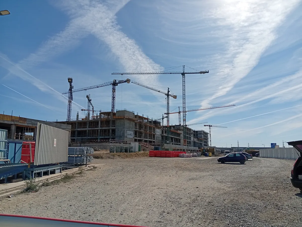 The building site of Craiova's new hospital, October 2026: a forest of tower cranes over concrete frames already four and five storeys high, with site huts and stacks of building blocks in the gravel yard in front