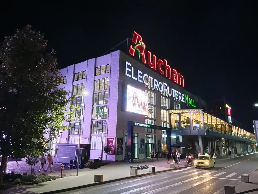 The frontage after dark — the Auchan sign in red neon over the old factory brick, the lit arcade running away down the side